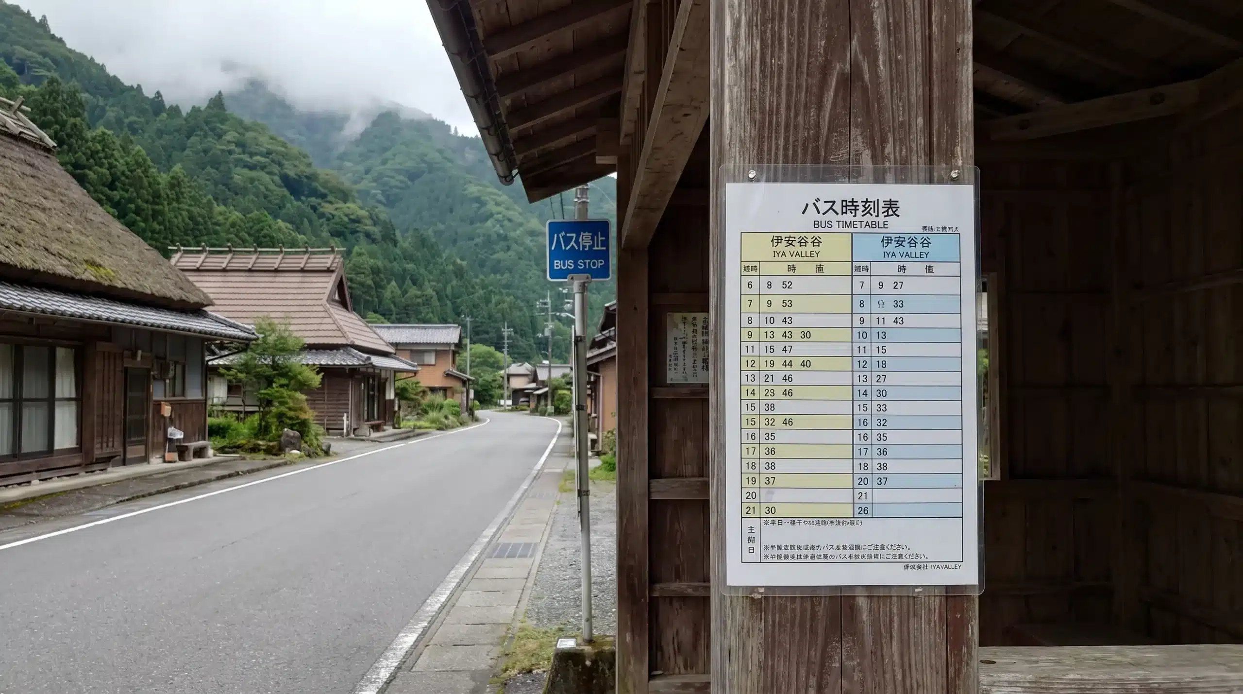 A Japanese-language timetable stands at a rural bus stop in the Iya Valley, beside a narrow road and traditional houses with mountain slopes behind them.